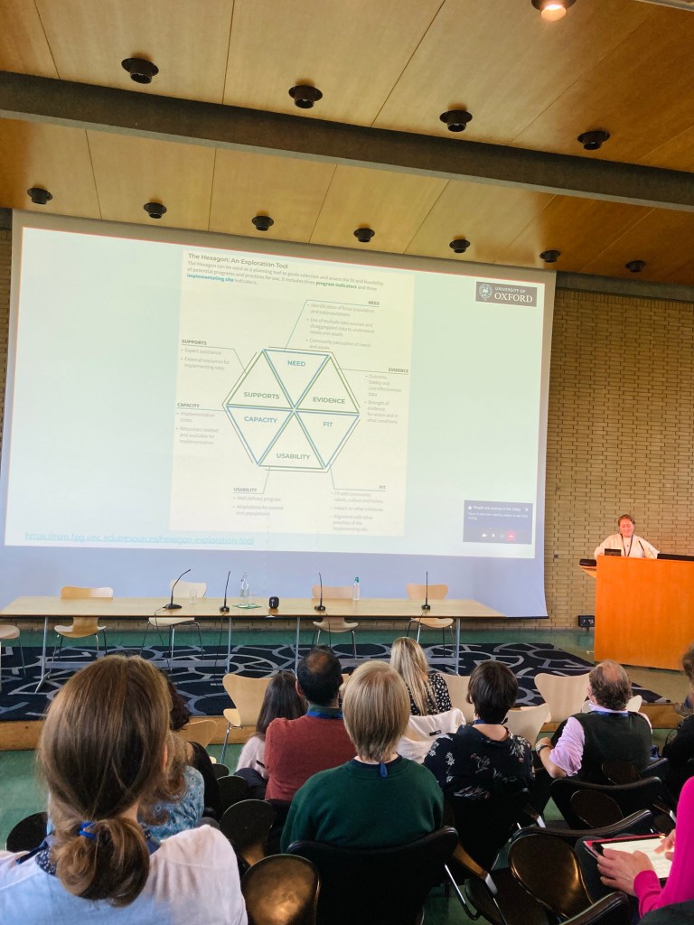 A woman presenting at a lectern next to a large screen in a lecture theatre, which shows the Hexagon (an exploration tool).