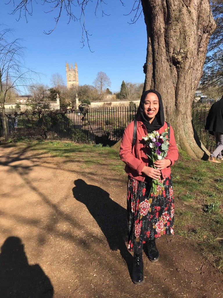 A woman wearing a black headscarf, red jacket and patterned skirt smiling and holding a bunch of flowers. She is standing under a tree in a meadow with a tall chapel tower visible in the distance. 