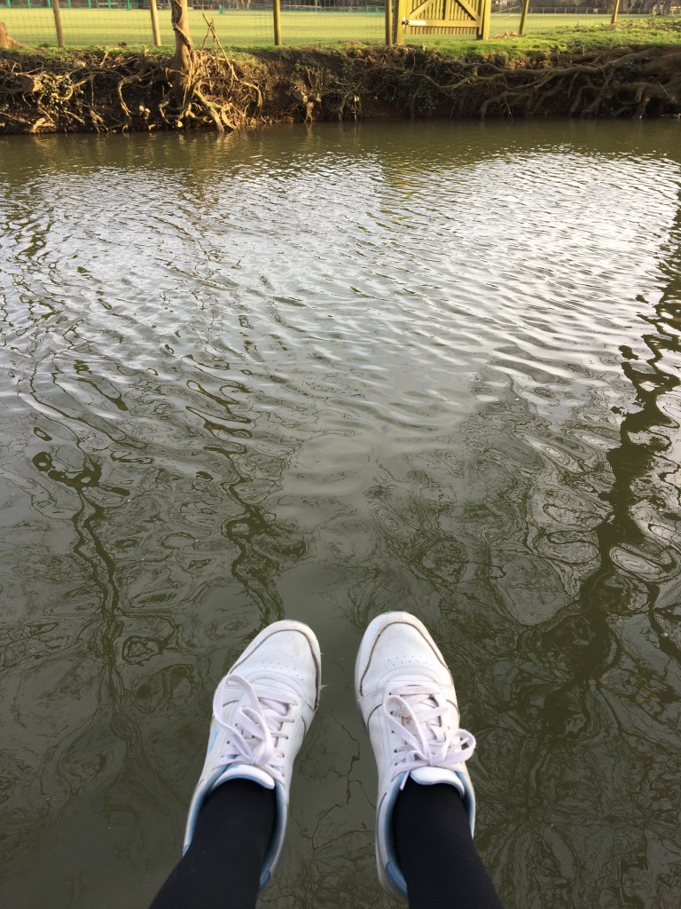 Murky brown river water with a field beyond the river bank. Two feet are visible dangling over the water, wearing white trainers and black tights.